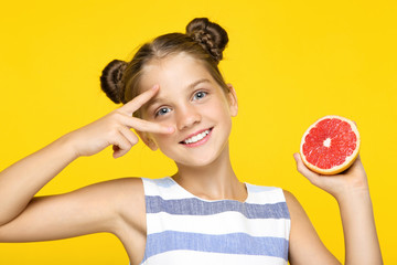 Wall Mural - Young girl holding grapefruit and showing two fingers on yellow background