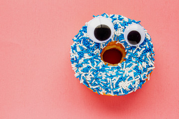 Wall Mural - Vanilla Dip Donut with Black Wiggle Googly Eyeballs on a Coloured blue and white Sprinkles on a pink background.
