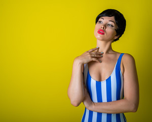 Wall Mural - Brunette in a striped swimsuit posing in the studio on a bright yellow background. Woman in a short wig with sensual lips.