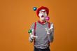 Mime in vest and red hat juggles with colorful balls on orange background