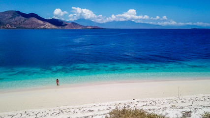 A drone shot of a girl playing on the beach on a small island near Maumere, Indonesia. Happy and careless moments. The coast changes colors from white to turquoise and navy blue. There are few islands