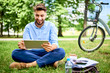 © baranq - Smiling young man sitting in park with tablet and bicycle during lunch break