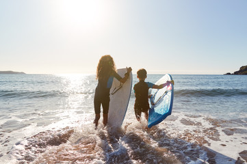 Rear View Of Two Children Wearing Wetsuits Running Into Sea With Bodyboards On Beach Vacation