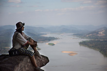 Asian Thai Woman Travel Visit And Posing Sitting On Ridge Stone Of Cliffs At Wat Pha Tak Suea Temple With View Of Landscape Of Nongkhai City And Loas And Mekong River In Nong Khai, Thailand