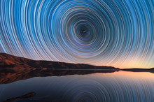 Beautiful Night Landscape With Small Lake And Mountains. The Colorful Star Trails On The Sky Reflacted On The Water. Night Time Lapse Photography.
