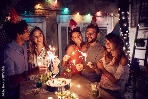Fotomural Smiling group of young friends having birthday party in the club with cake and candles at night