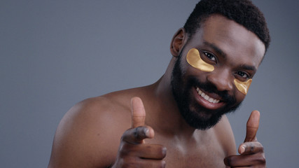 Portrait of cheerful gentle african young man applying collagen patches under the eyes pointing with forefingers smiling at camera.