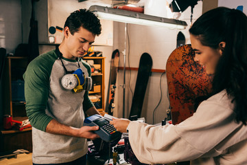 Wall Mural - worker holding terminal and smiling woman paying with credit card and holding snowboard in repair shop