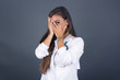 © Jihan - Beautiful young caucasian doctor woman covering her face with her hands and peering out with one eye between her fingers standing indoors. Scared from something or someone.