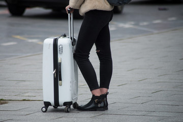 Closeup of legs of girl wearing black jeans and black shoes waiting with white suitcase in the street