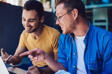 Smiling prosperous male it professionals pointing on laptop computer web page satisfied with cooperation on project, happy hipster guys laughing on social networks video talking to each other.