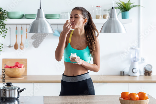 Sporty young woman eating iogurt while standing in the kitchen at home.