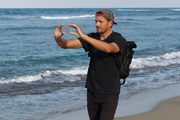 Man tourist with backpack using cell phone to take photo, standing on a beach.