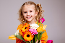 Little Blonde Girl In An Orange Dress Holds Tulips And Gerberas In Her Hands On A White Background, A Child Girl Smiles And Holds Spring Flowers In Her Hands, Space For Text