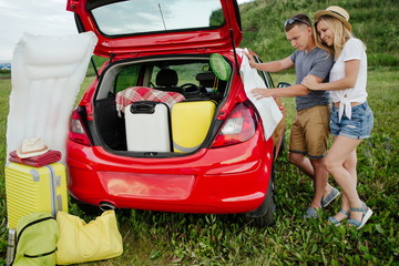 Wall Mural - Young married couple looking at a map of the area, camping in nature. Red car loaded with Luggage for travel