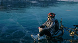 Fototapeta Do akwarium - Woman sitting near her bike on ice. The girl cyclist stopped to rest. She sits on the wheel and enjoys the beautiful view of the sunset. The traveler is ride a cycle. Female is riding bike on the ice.