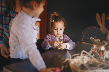 Two toddlers, sisters, with mother, making Easter decorations in their living room.