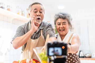 Couple senior Asian elder happy living in home kitchen. Grandfather cooking salad dish with grandmother recording vlog vdo for social blogger.  Modern lifestyle older people relationship and activity.