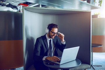 Smiling handsome caucasian bearded businessman in suit, with eyeglasses and headphones sitting at workplace and typing on laptop.