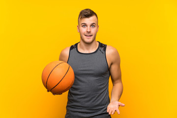 Young handsome blonde man holding a basket ball over isolated yellow background with shocked facial expression