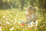 Fototapeta Tulipany - Portrait of happy mother and her little child on spring background. Cheerful family at dandelion field.Mom and her cute daughter outdoors.