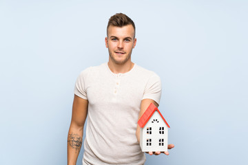 Wall Mural - Young handsome blonde man over isolated blue background holding a little house