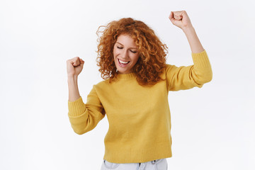 Carefree, attractive modern redhead girl in yellow sweater, dancing as triumphing feeling joy of success, delighted, moving hands, fist pump, close eyes and smiling pleased, white background