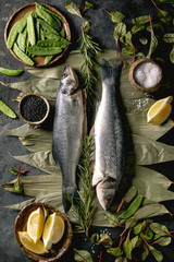 Wall Mural - Raw fresh uncooked fish sea bass on dry bamboo leaves ready to cook with lemon, sea salt, black and green beans, green salad over dark texture concrete background. Flat lay, space