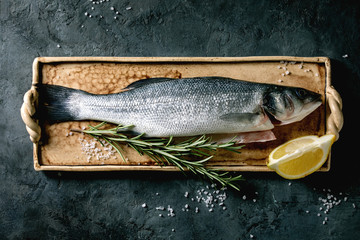 Wall Mural - Raw fresh uncooked fish sea bass on ceramit plate ready to cook with lemon, sea salt and rosemary over dark texture concrete background. Flat lay, space