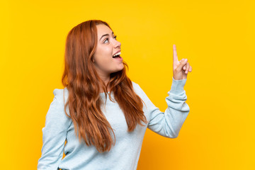 Wall Mural - teenager redhead girl over isolated yellow background pointing up and surprised