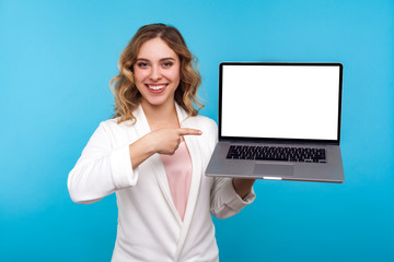 Look at internet advertisement. Portrait of happy businesswoman with wavy hair in white jacket pointing laptop with blank screen and smiling at camera. indoor studio shot isolated on blue background