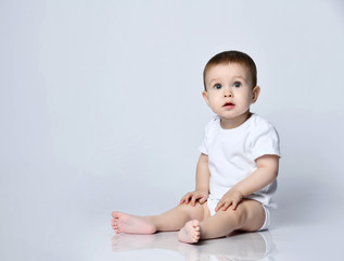 infant baby boy toddler in white bodysuit is sitting sideways on the floor looking up at us on a gra