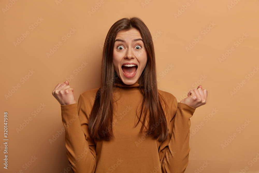 Portrait of emotive cheering woman with European appearance, clenches fists, finds out about her win, clenches fists with triumph, shares success, yells I am winner, poses over beige studio wall - obrazy, fototapety, plakaty 