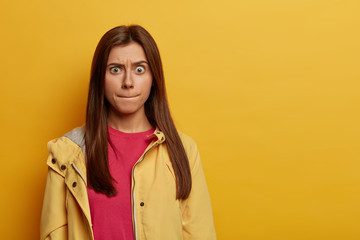 Wall Mural - Portrait of emotional worried European woman presses lips, tries to control her emotions, raises eyebrows, wears pink jumper and anorak, stands indoor over yellow background, copy space aside