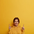 Friendly looking lovely woman shows promo with joy, points above with both index fingers, gives recommendation or advice, wears yellow hoodie, found awesome promo, suggests nice place upwards