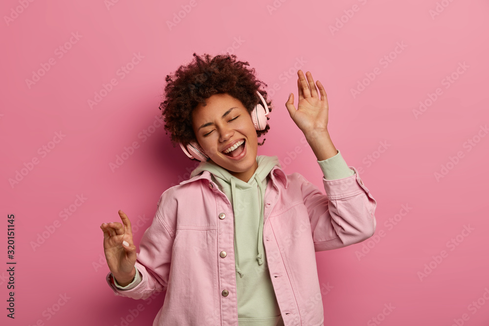 Joyful curly haired teenage girl dances carefree listens audio track in headphones, enjoys playlist, sings song along, wears casual apparel, poses against pink pastel background, expresses happiness - obrazy, fototapety, plakaty 