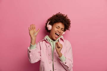 Wall Mural - Photo of overjoyed woman sings along in headphones, makes microphone with hand, raises palm and dances carefree, being in positive mood, moves with rhythm of music, wears fashionable clothes