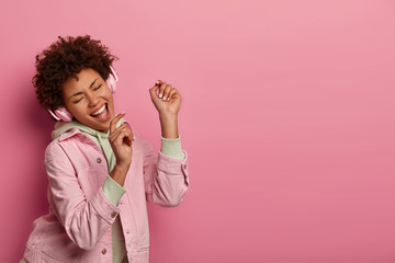 Portrait of joyful expressive ethnic woman holds hands like microphone, enjoys favourite track in headphones, raises palms and dances happily, dressed in casual clothes copy space aside for your promo