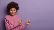 Curious pleased curly girlfriend points at upper right corner with both index fingers, asks boyfriend for hang out, wears casual rosy jumper, isolated over purple background, shows awesome promo
