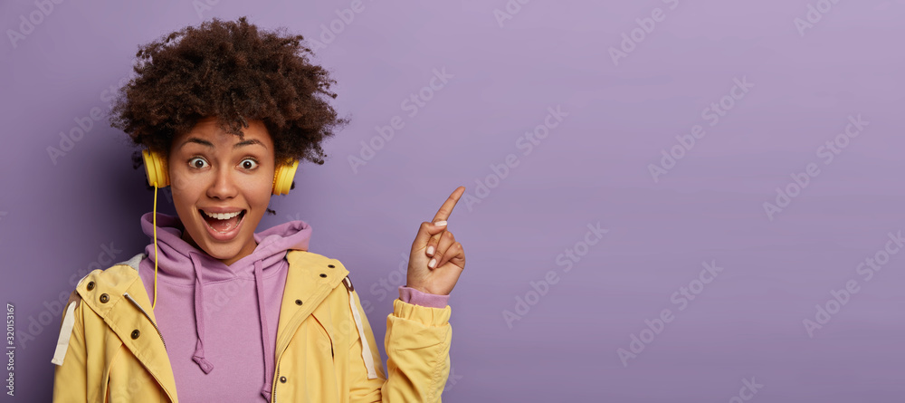 Millennial hipster girl with Afro hair enjoys music podcast in headphones, points fore finger on copy space for your advertising, has excited surprised happy expression, isolated on purple background - obrazy, fototapety, plakaty 