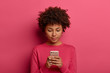© WHstudio Leushin N - Photo of serious curly woman uses modern device, orders taxi via smartphone application, texts or browses in social media, wears bright crimson clothes, isolated on pink background, checks email box