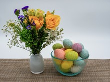 Flower Arrangement In A White Case With A Glass Bowl Of Decorated Pastel Colorful Easter Eggs On Top Of A Beige Patterned Table Runner With A White Background.
