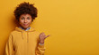 Recommend you to use copy space. Calm lovely Afro American woman points thumb aside, shows nice place to visit, wears hoodie, isolated on yellow studio background, suggets good offer or proposal