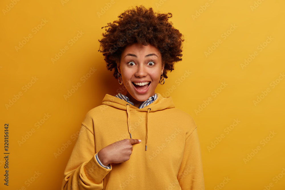 Joyful teenage girl points at herself, happy to be chosen to participate in international project, laughs happily, wears casual yellow sweatshirt, asks who me, cannot believe in her unexpected success - obrazy, fototapety, plakaty 