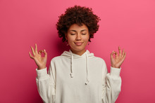 Stress Goes Away. Relaxed Peaceful Woman Raises Hands In Zen Gesture, Closes Eyes And Practices Yoga, Tries Stay Calm, Concentrated On Something, Wears White Hoodie, Isolated On Vivid Pink Background
