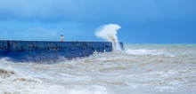 A Harbour Wall With A Rough Sea Crashing Against It And A White Wave Crashing 20 Meters Into The Airand A Lighthouse In The Background, 