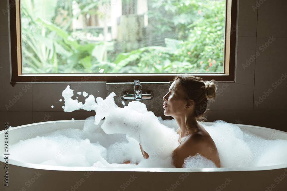Woman relaxing in foam bath with bubbles in dark bathroom by window - obrazy, fototapety, plakaty 
