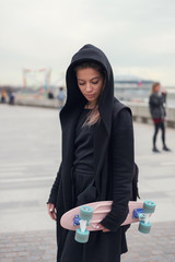 Portrait of a skater style teenager girl in black hood-jacket with pink skateboard