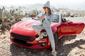 Wall Mural - Young woman dressed casually with hat enjoying road trip on the island, standing with phone near the convertible car on the roadside