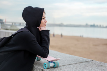 Portrait of a skater style teenager girl in black hood-jacket with pink skateboard